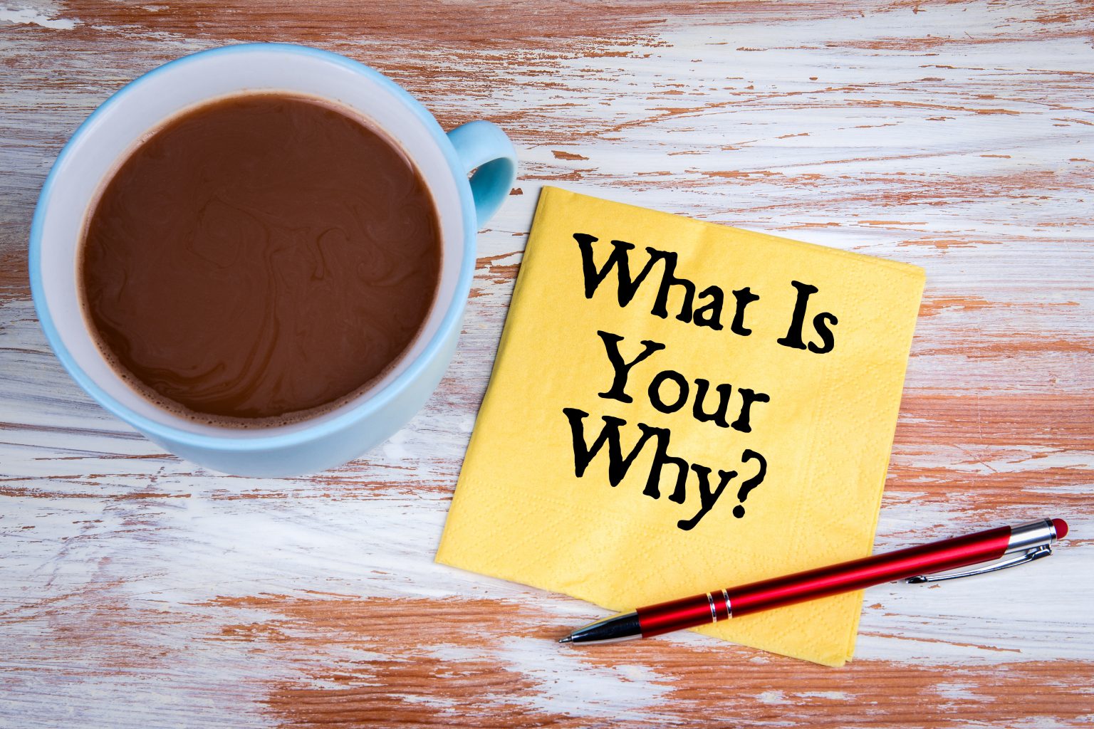 A cup of hot chocolate next to a yellow napkin with "what is your why?" printed on it, accompanied by a red pen, all set on a wooden surface.