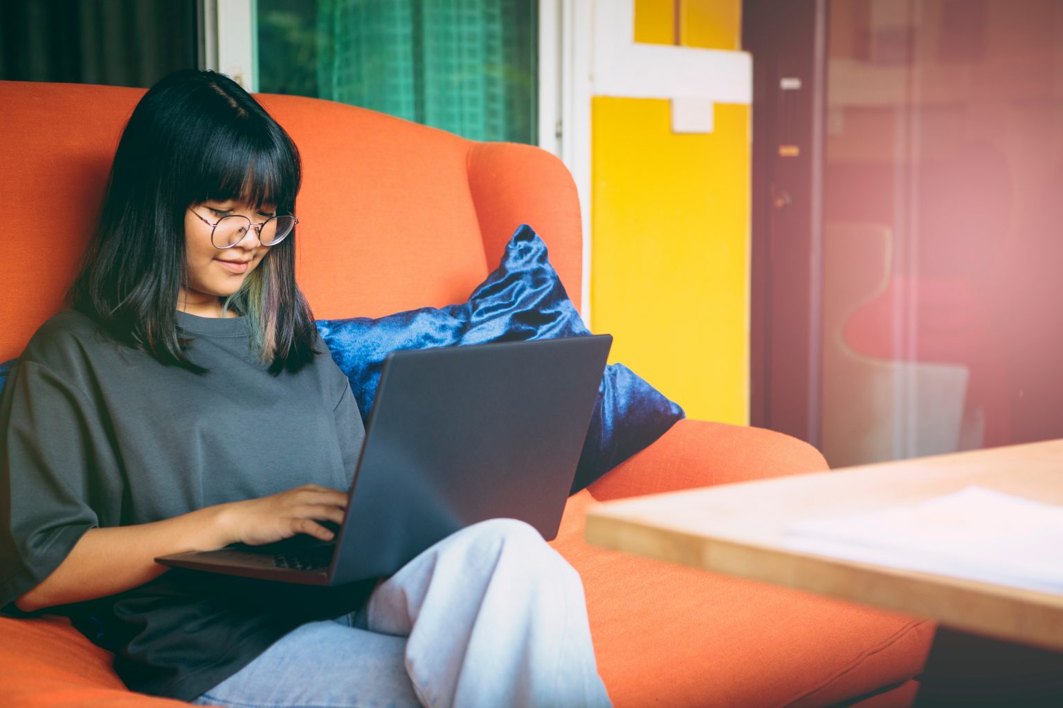 asian young woman working on laptop computer while sitting on couch in cheery bright room